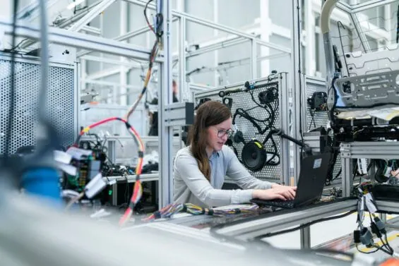 A woman works on a laptop as she sits in a workspace surrounded by wires and tech equipment.