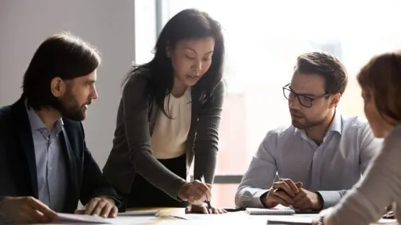 A woman explains a concept while referencing a piece of paper to young professionals sitting at a table together.