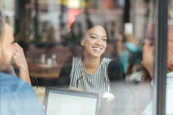 A young woman seen through a glass window smiles at her colleagues as they work together at a table.