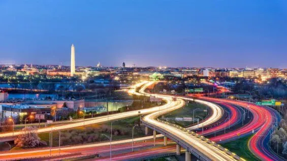 A brightly lit highway passes over a river at dusk with the Lincoln Memorial visible in the background.