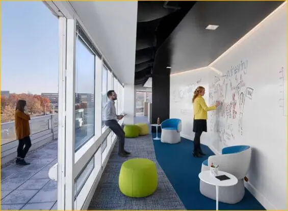 A woman writes at a whiteboard as a coworker observes her in a modern office building, while another coworker is visible outside on the balcony.