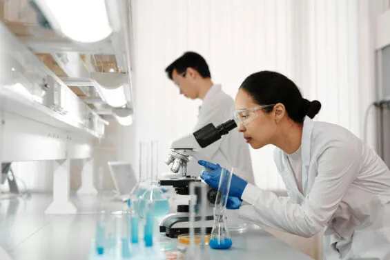 A scientist in a lab coat and protective gear uses a microscope in a lab.