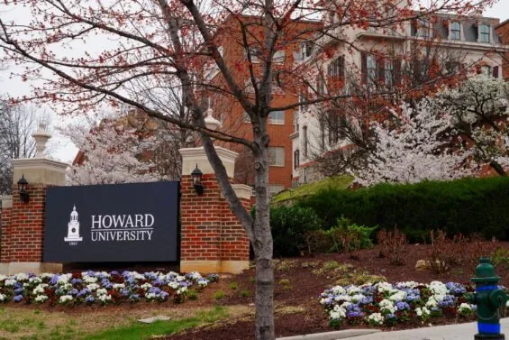 A sign on a landscaped slope reads Howard University with brick buildings and flowering trees in the background.