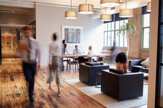 A modern coworking space features people working at a large table or sitting in comfortable chairs as others walk by.