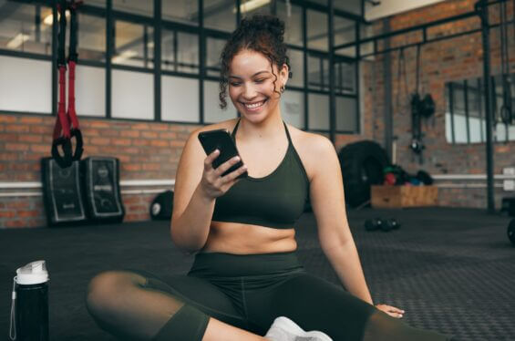 A young woman in exercise clothing smiles at her phone while sitting on a yoga mat at a gym.