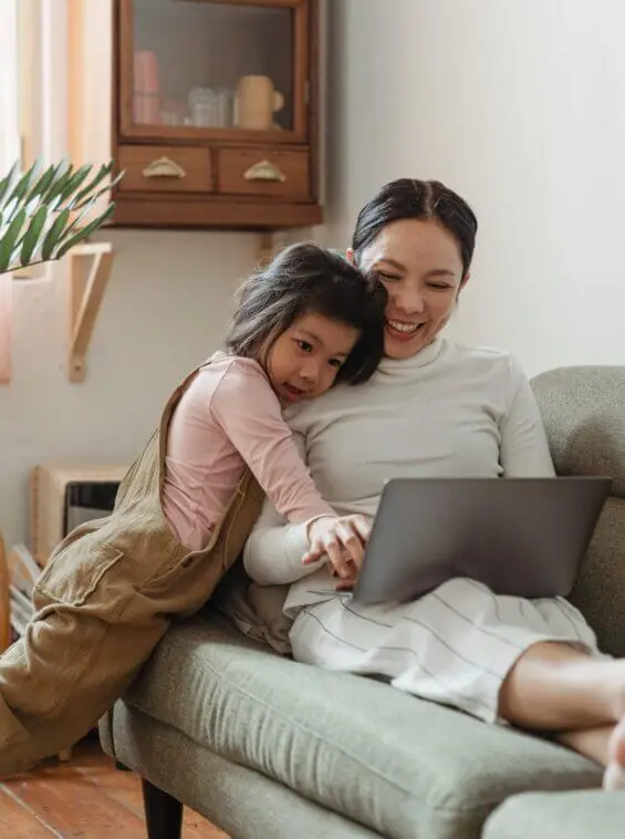 A young woman sits on a couch and smiles as she uses her laptop with her daughter next to her.