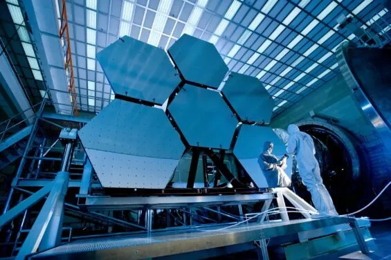 Two workers in protective suits stand next to large mirrored equipment.