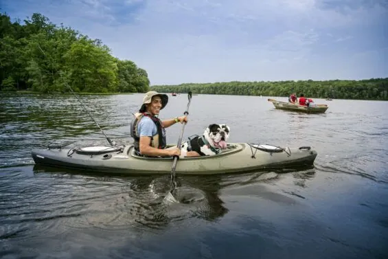 A young man smiles while paddling with his dog in a canoe on a river.