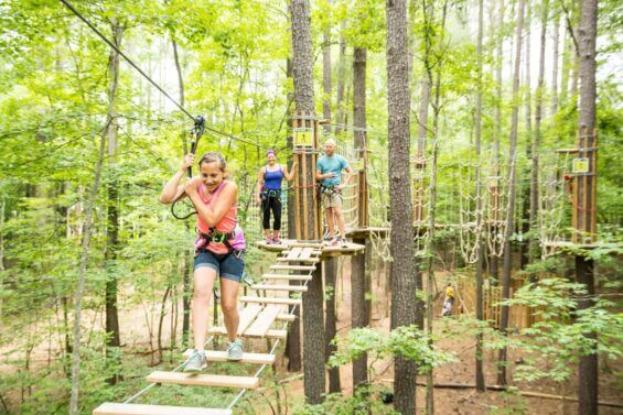 A young girl with a safety harness walks across an adventure obstacle course while her parents watch in the background.